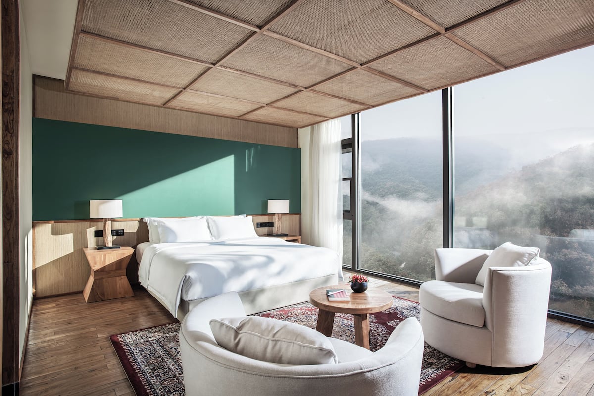 Una camera d'albergo moderna con un letto grande, due comodini, un divano, un tavolino rotondo e un'ampia finestra con vista sulle montagne.