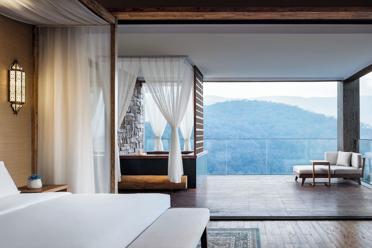 Una camera da letto con un letto grande, un comodino, una lampada e vista su un balcone con una sedia.