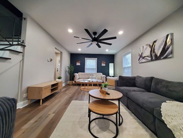 A modern living room with a dark grey sofa, a wooden coffee table, and a ceiling fan. - Modern Oasis in Glen Ellyn (Glen Ellyn)