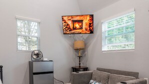 Uma sala de estar com um sofá seccional cinza, uma mesa pequena, um abajur e uma TV fixada na parede.