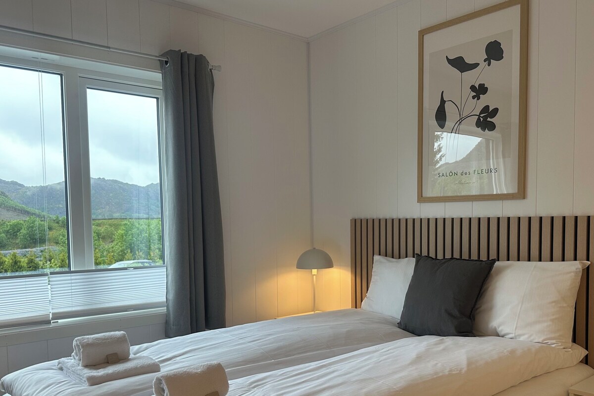 Una habitación de hotel con una cama grande, una lámpara de noche, un cuadro enmarcado en la pared y una ventana con cortinas.
