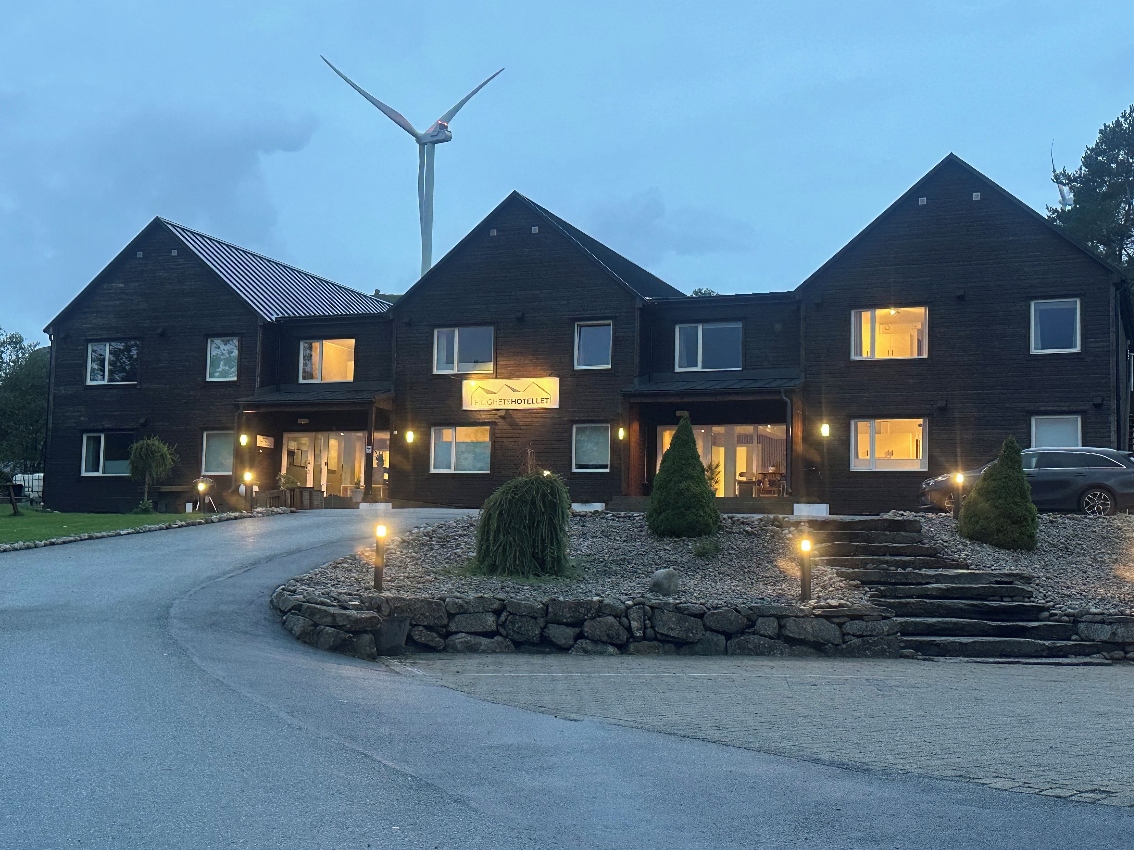Un hôtel à deux étages, doté d’une façade sombre, avec une éolienne en arrière-plan et une entrée bien éclairée.