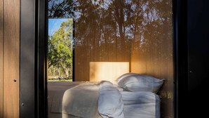 Un letto con lenzuola e cuscini bianchi, una testiera in legno e vista su alberi attraverso una finestra.