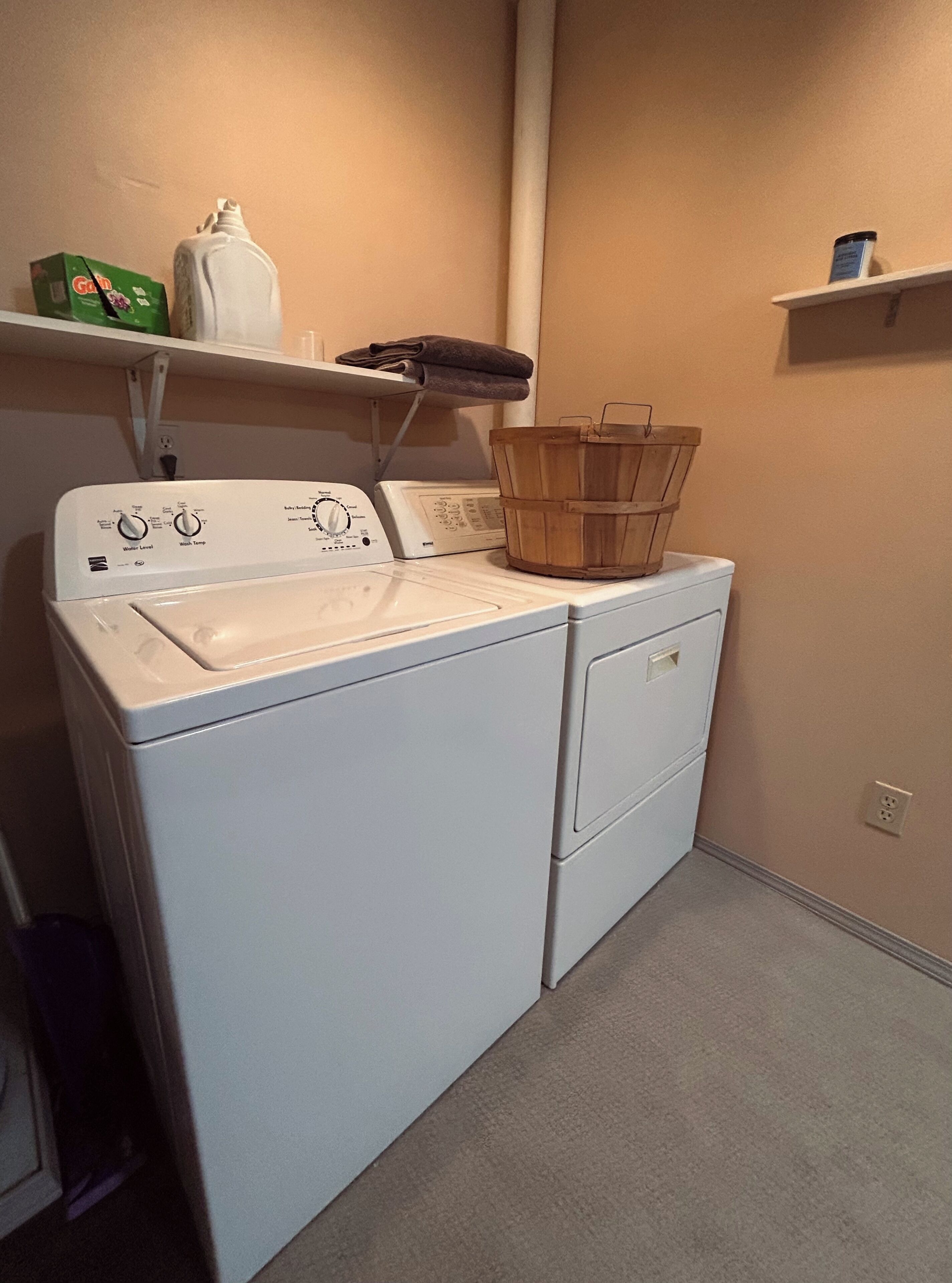 A compact laundry area with a front-loading washer and dryer, a wooden laundry basket on the dryer, and shelves above holding a bottle and a box.