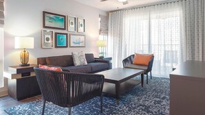 Een moderne hotelkamer met een bank, fauteuil en een bijzettafeltje. Er hangen afbeeldingen in lijsten aan de muur, een lamp en een groot raam met gordijnen.