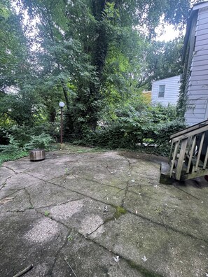 A concrete patio with a metal grill, surrounded by dense foliage and a wooden deck.