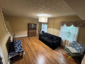 Quarto com piso de madeira, um sofá preto, uma mesinha, um armário e uma janela com cortinas.