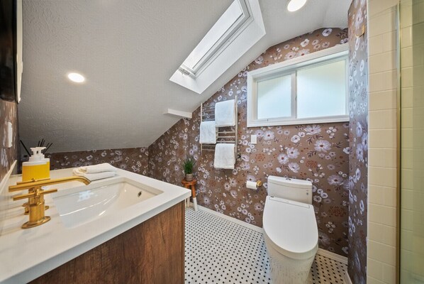 A bathroom with a white sink, gold faucet, and a toilet. There is a window above the toilet and a shower curtain on the right.
