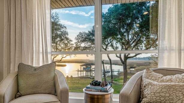 A room with two beige armchairs facing a large window overlooking a lake and trees. A small round table with a book titled "Bird Songs" is in the center. - Florida Riverfront Sunrise | Fish, Boat, Unwind (Jacksonville)