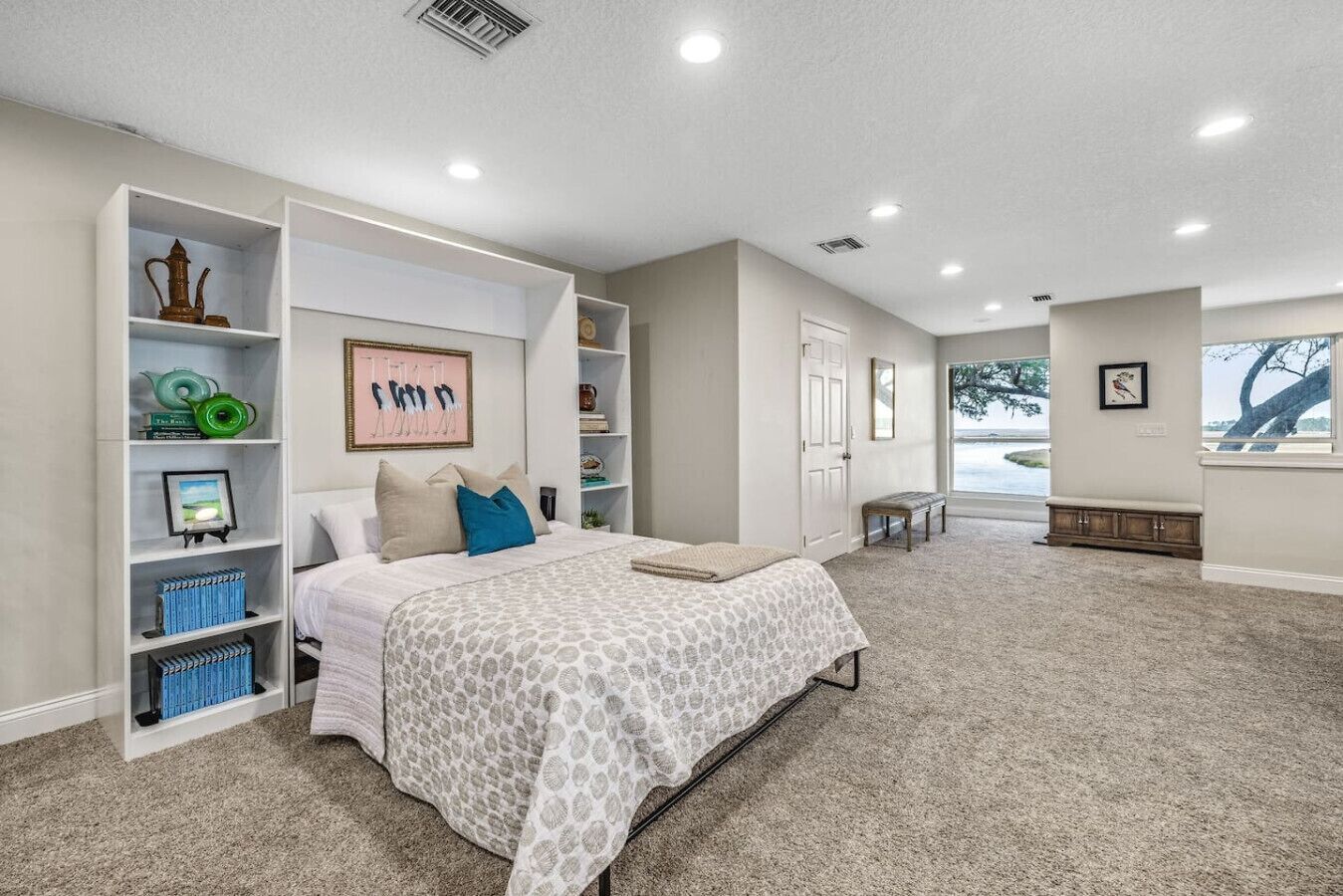 Un dormitorio con una cama grande, estanterías empotradas con libros y objetos decorativos, un banco y vistas al exterior.