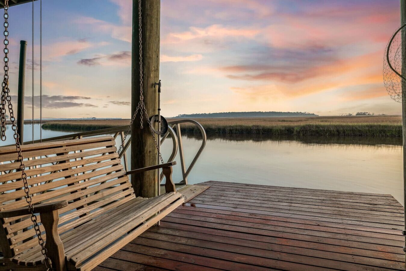 A wooden deck with a bench and a swing, overlooking a calm body of water and a distant shoreline.
