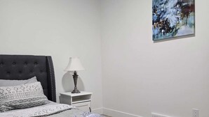 Un dormitorio con cama, mesita de noche, lámpara, un cuadro en la pared y una rejilla de ventilación.