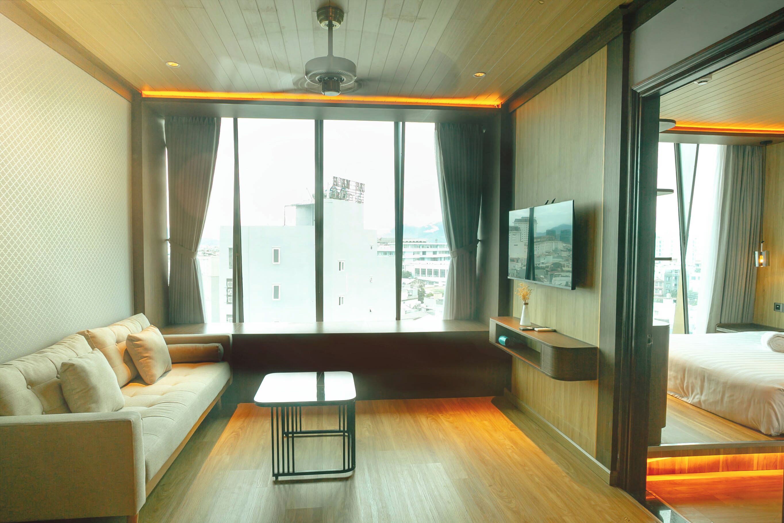 Hotelkamer met een bank, een kleintje tafeltje, een bed, een televisie en grote ramen met gordijnen.