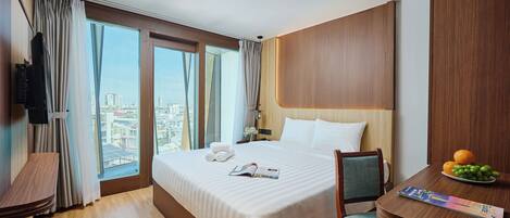 A hotel room with a large bed, a desk, and a chair. There is a TV mounted on the wall, and a window with curtains.