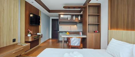 A modern hotel room with a large bed, a desk with a chair, a kitchenette, and a TV mounted on the wall.