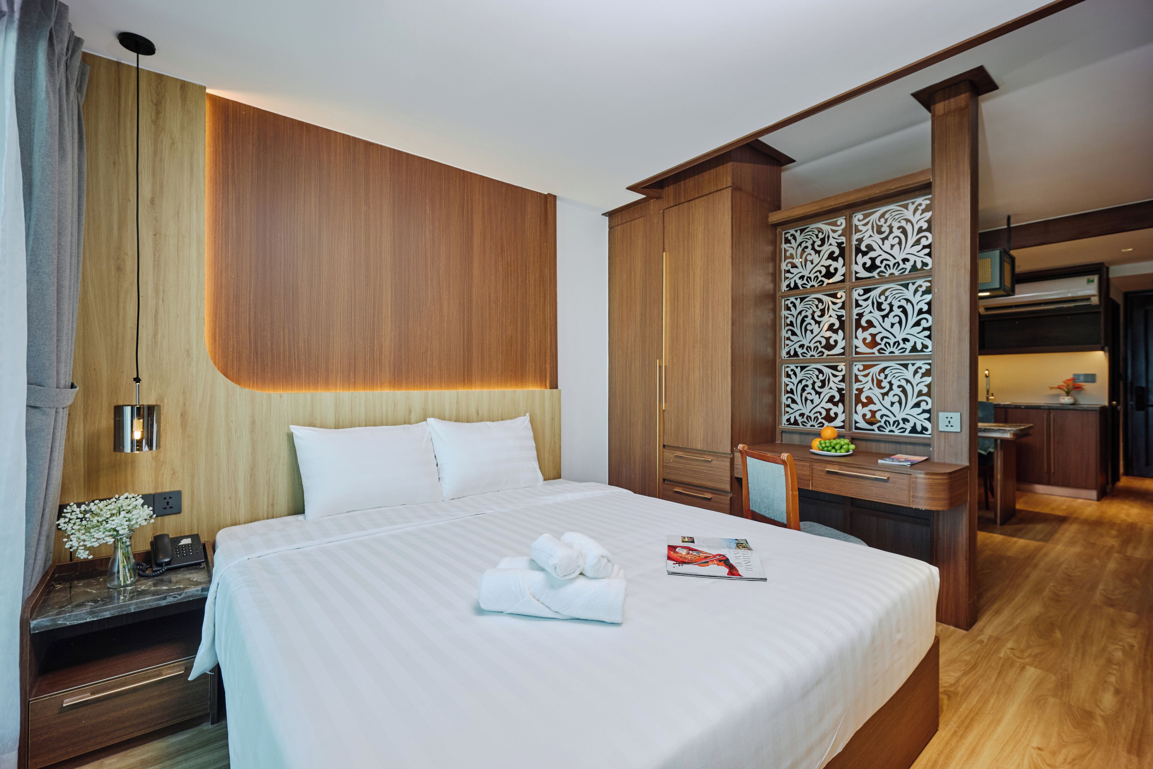 A hotel room with a large bed, wooden headboard, a desk with a chair, a small table with a vase of flowers, and a decorative panel with intricate designs.