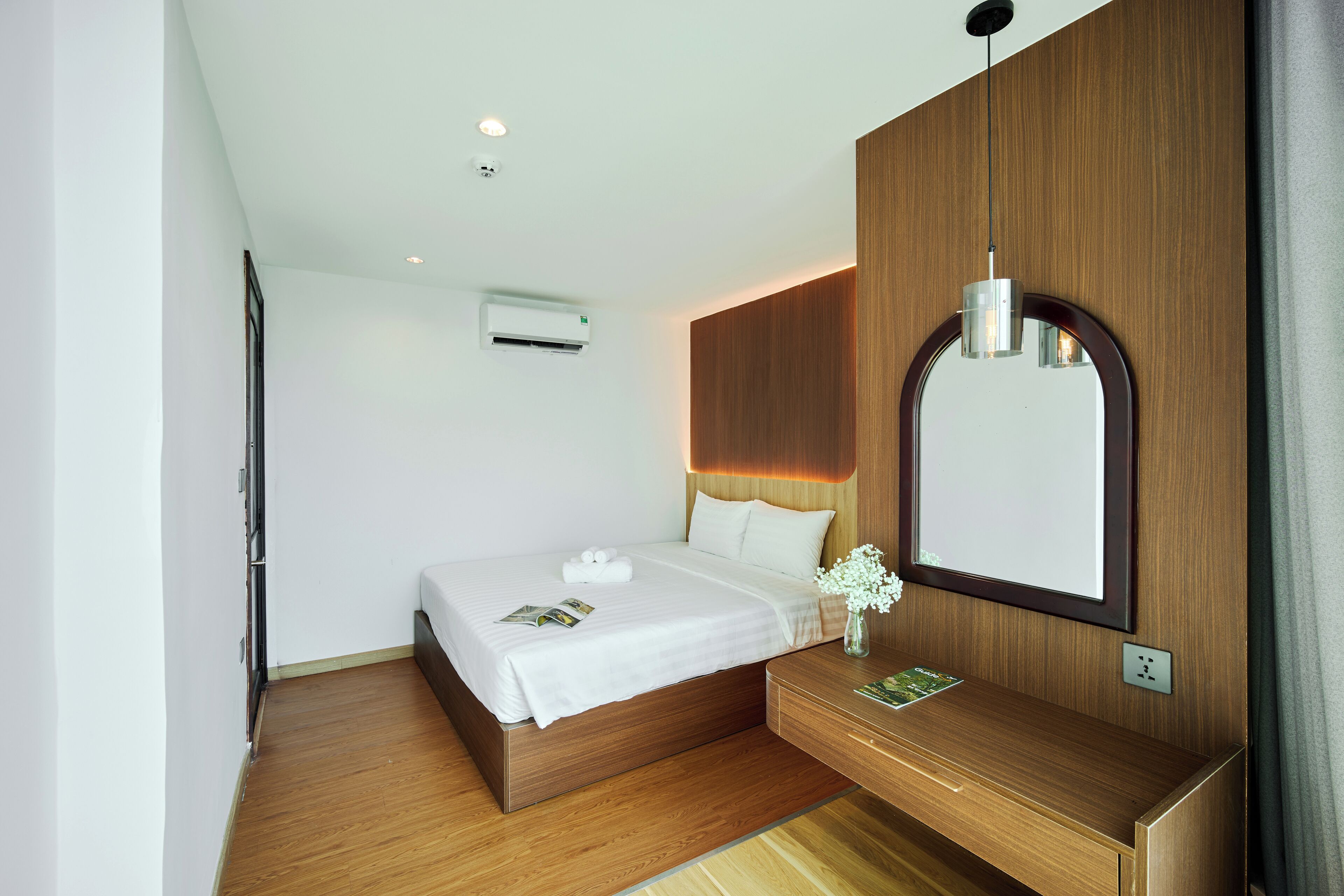 A modern hotel room with a wooden bed, a bedside table, a mirror, and an air conditioning unit.