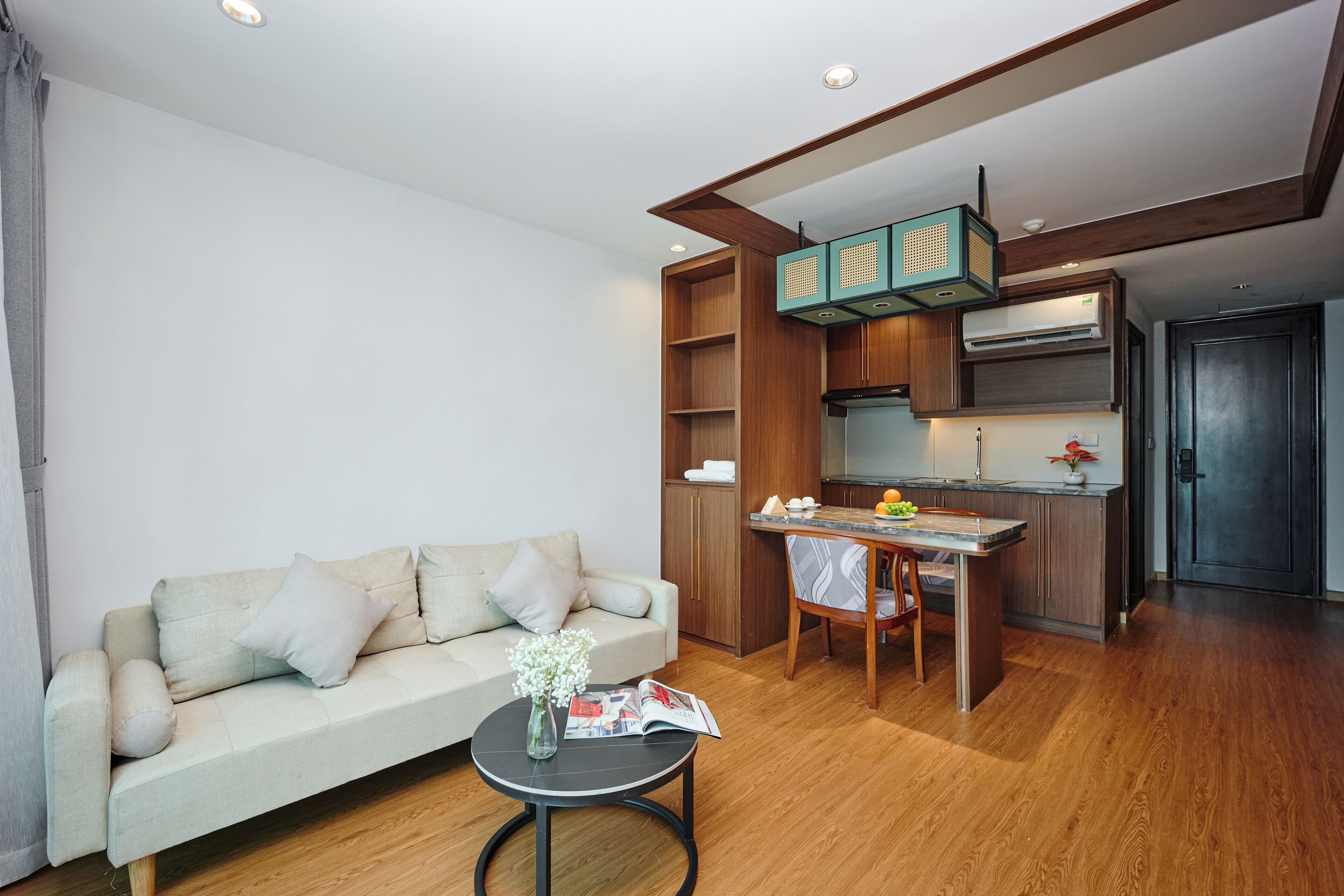 En kompakt opholdsstue med en sofa, et lille rundt bord med en vase og et magasin, et tekøkken med spiseplads, og en mørk dør.