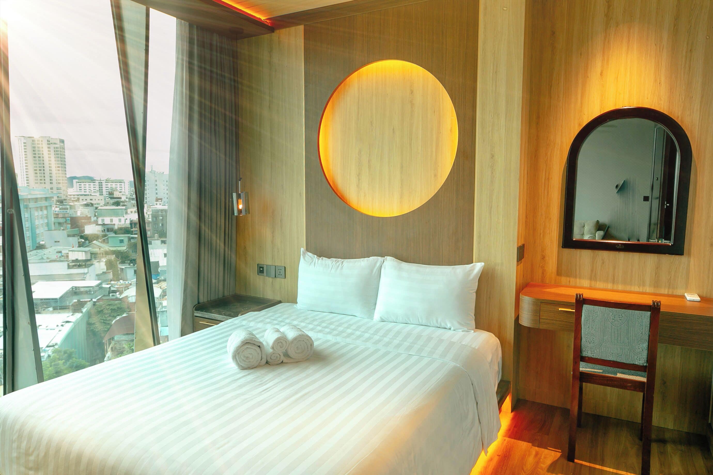 A hotel room with a large bed, a round wall-mounted light, a mirror, and a view of the city through a large window.