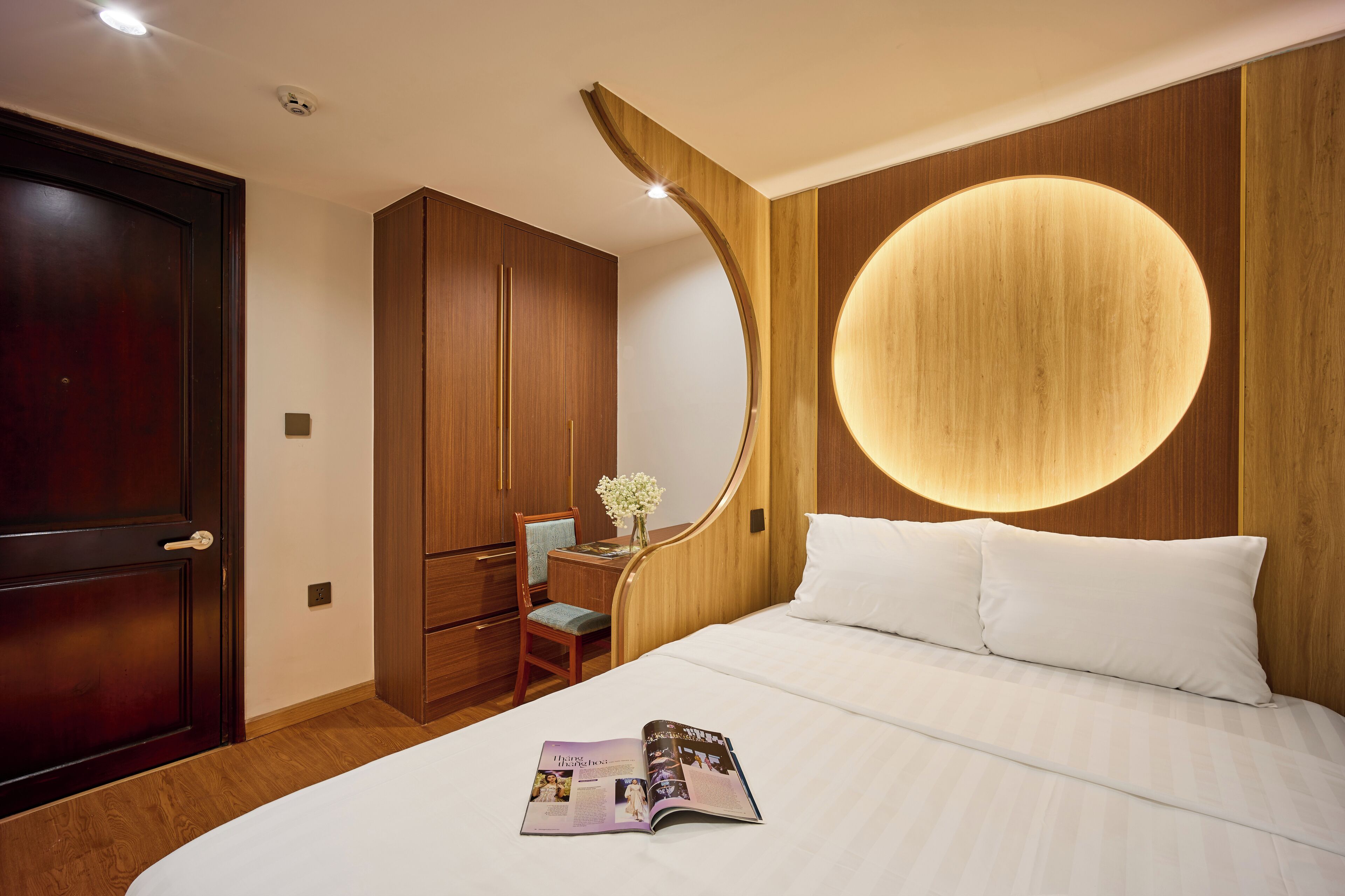 A hotel room with a large bed, a wooden wardrobe, a desk with a chair, and a round wall light.