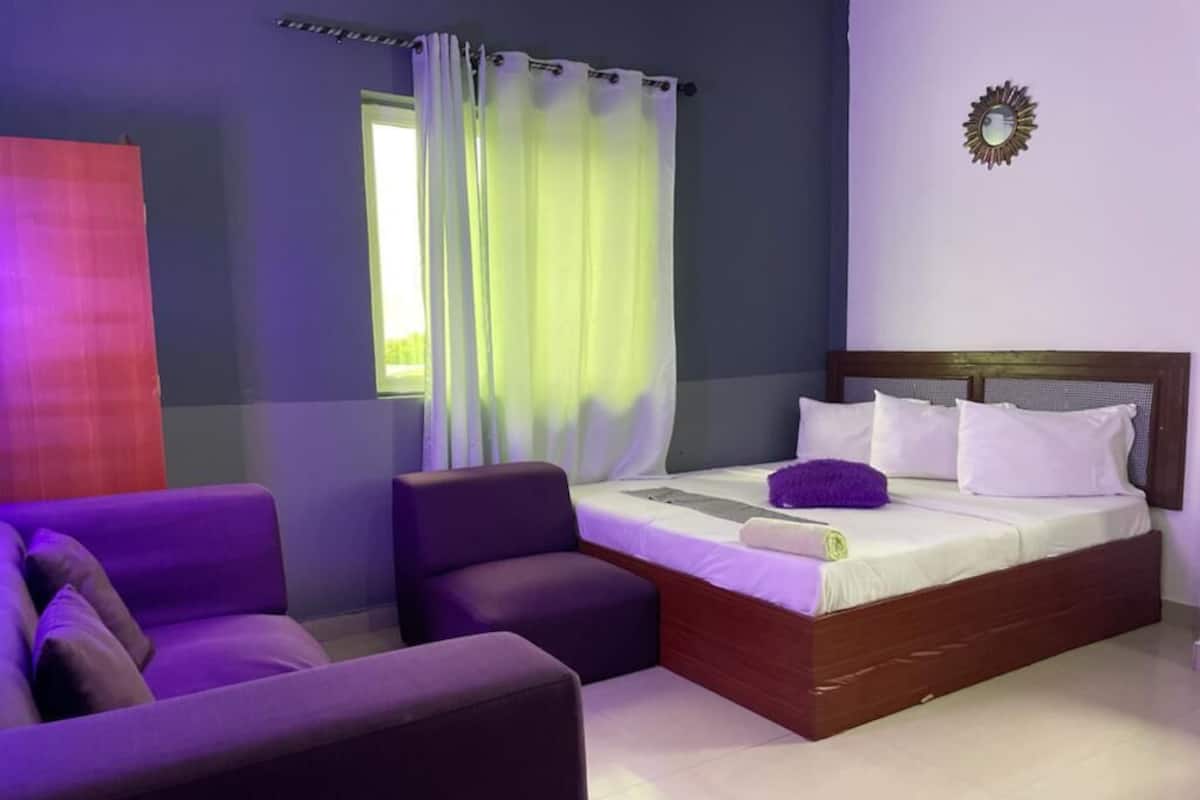 A bedroom with a bed, two purple chairs, a window with curtains, and a wall clock.