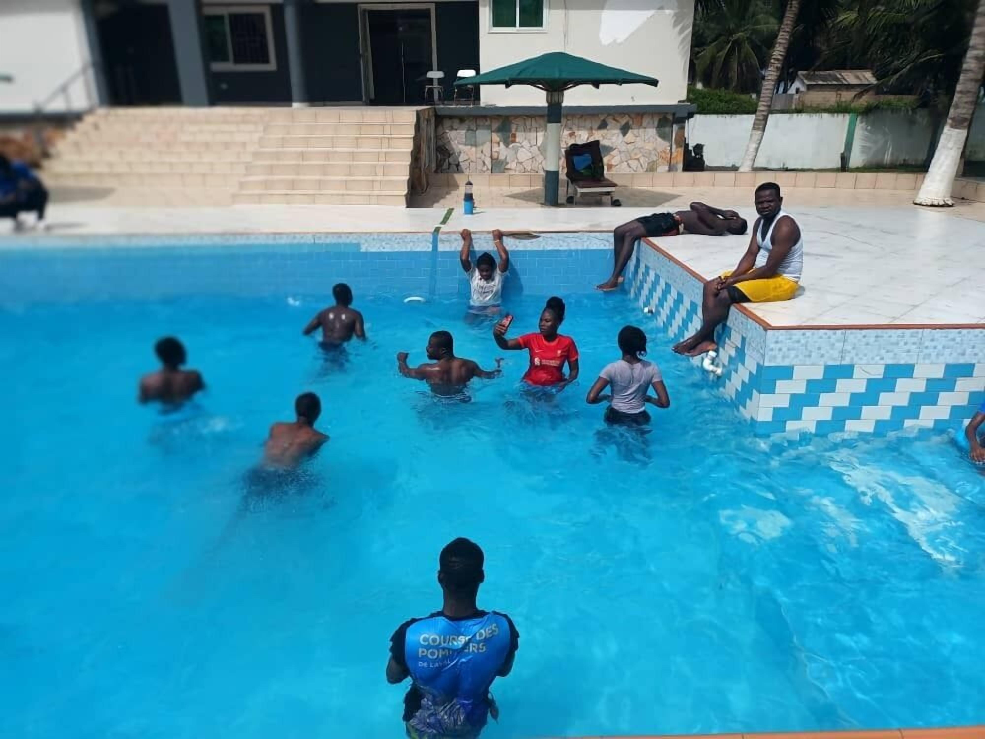 A swimming pool with people in and around it, a tiled deck, and a building in the background.