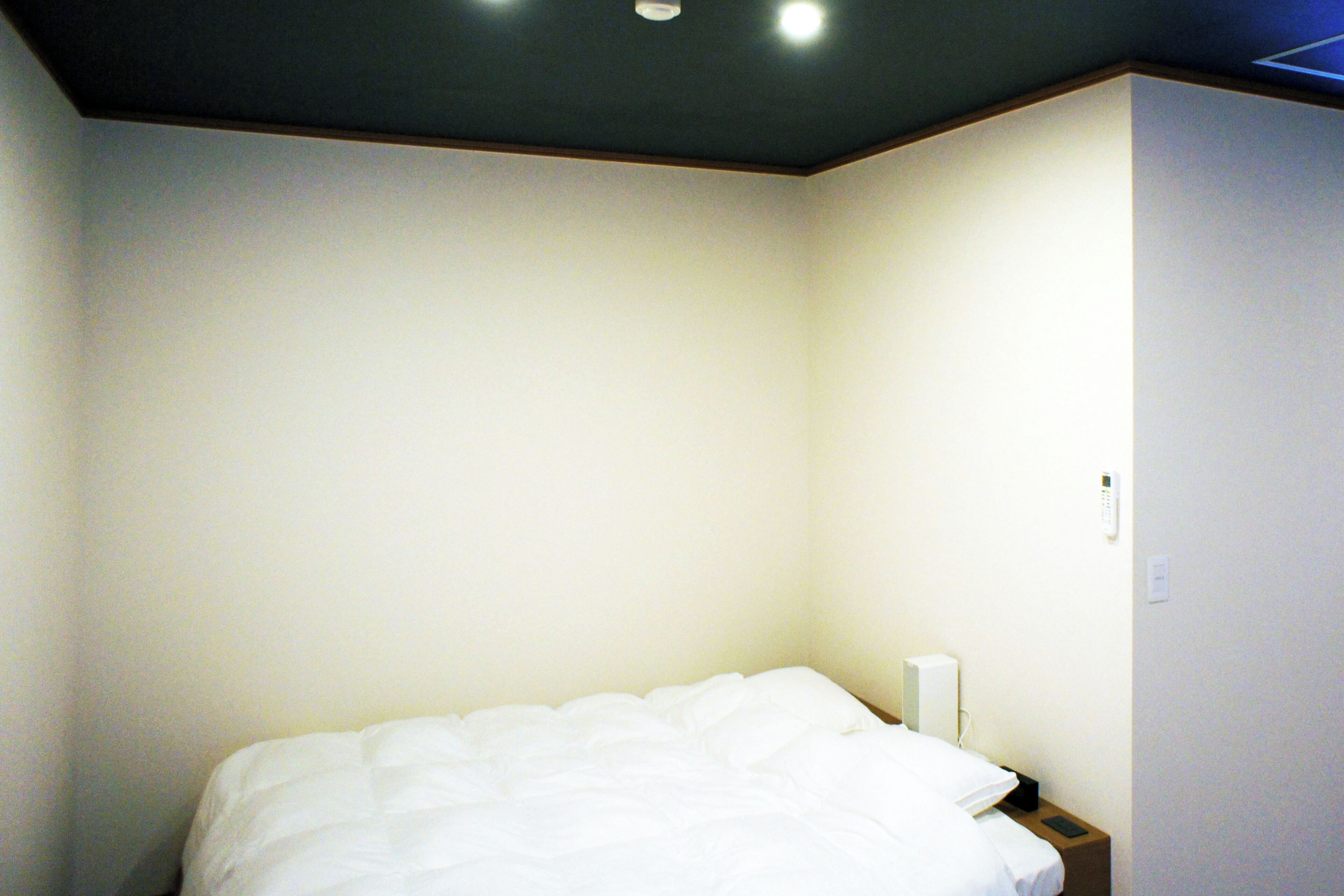 A simple, unadorned hotel room with a single bed, a bedside table, and a wall-mounted light fixture.