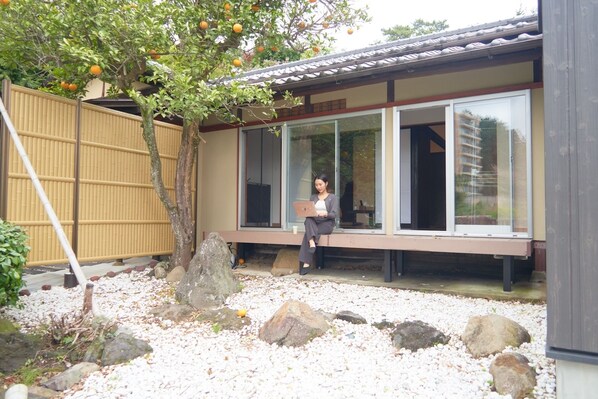 A person sitting on a bench outside a building with large windows, a tree with oranges, and a bamboo fence. - ATAMI SPRING TERRACE HOTEL (Atami)