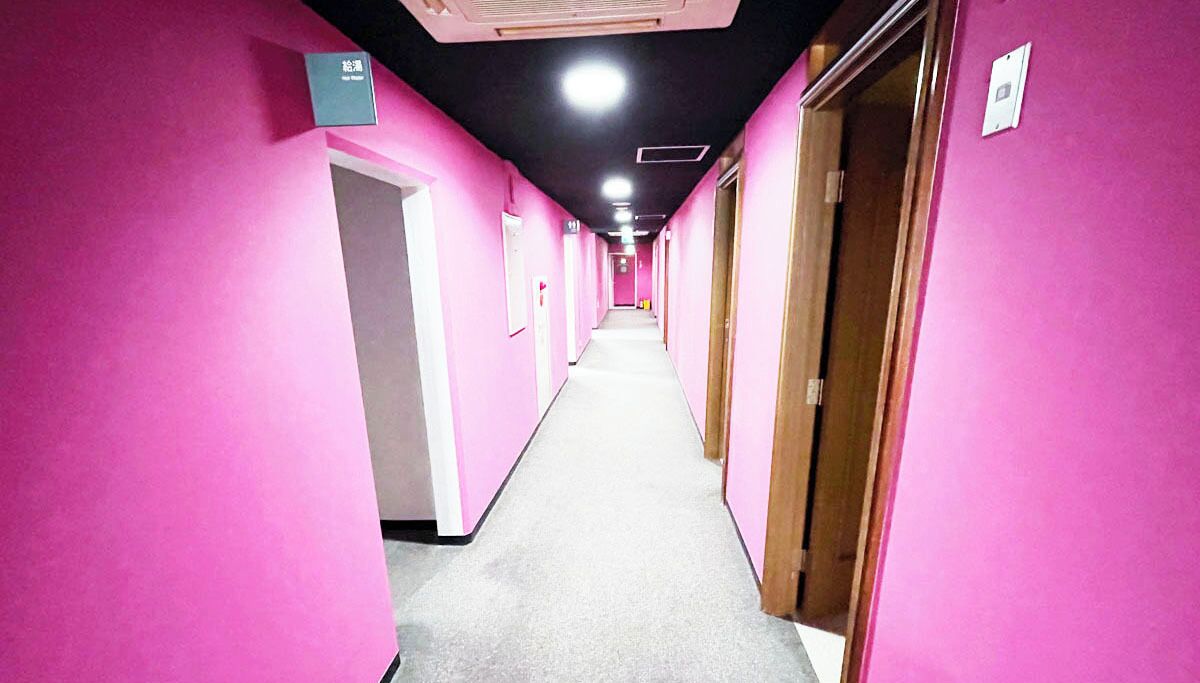 Un long couloir étroit aux murs roses et au sol recouvert de moquette, qui mène à plusieurs portes.