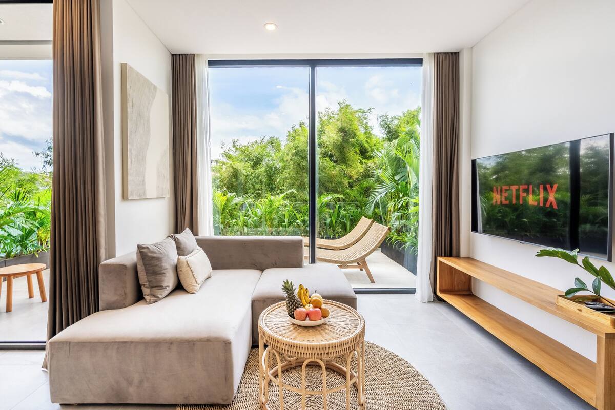 A modern living room with a sofa, a round table with fruit, a TV, and a view of greenery outside.