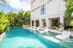 A swimming pool with white lounge chairs, surrounded by lush greenery and a modern building with large windows.