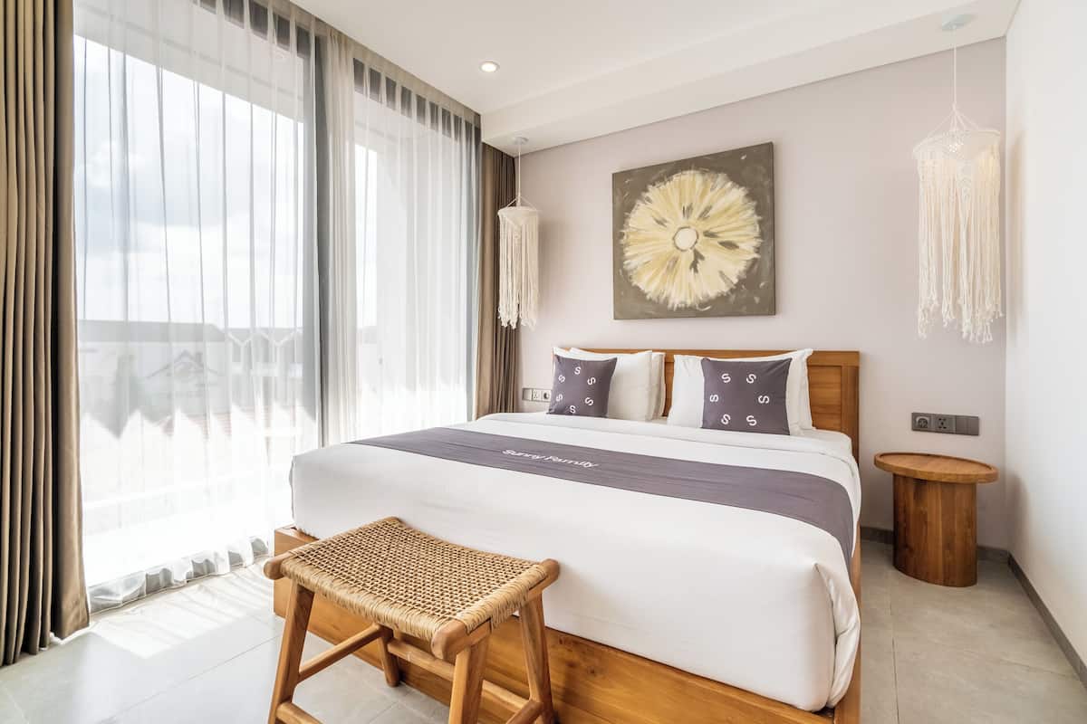 A modern hotel room with a large bed, a small wooden stool, a wall-mounted macrame light, and a large abstract painting.