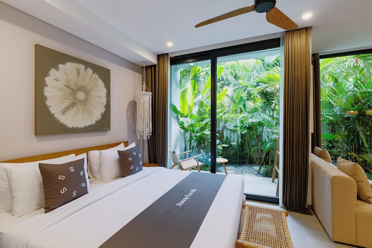 A modern hotel room with a large bed, a ceiling fan, a view of lush greenery through the glass door, and a seating area with a chair and table.