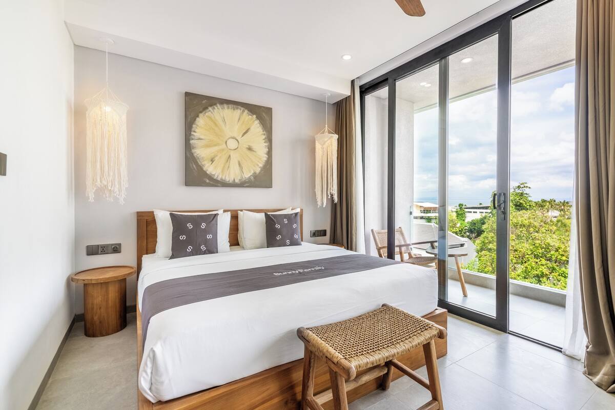 A modern hotel room with a large bed, a view of the outdoors, and a balcony with a table and chairs.