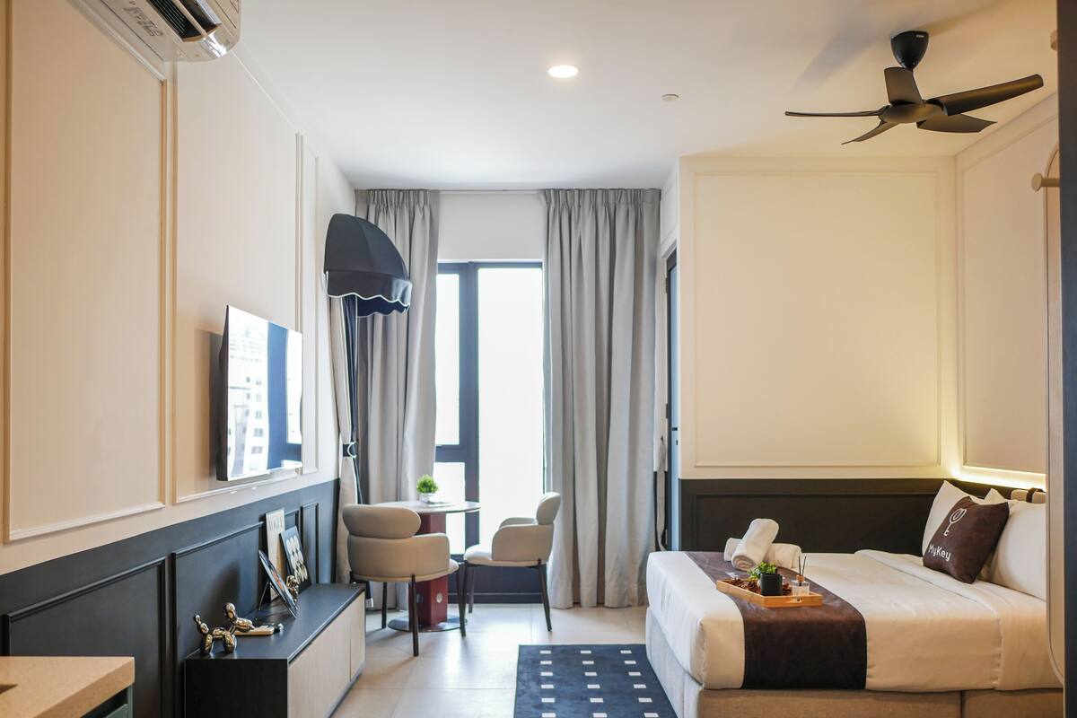 Ein modernes Hotelzimmer mit einem Bett, einem Schreibtisch, einem Sessel, einem Fernseher, einem Deckenventilator und einem Fenster mit Vorhängen.