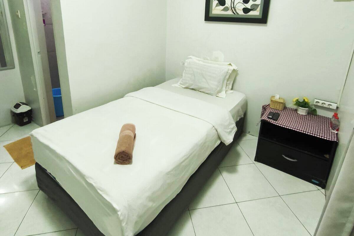 A single bed with a white mattress and a rolled towel on top, a small bedside table with a checkered cloth, and a wall-mounted light fixture.