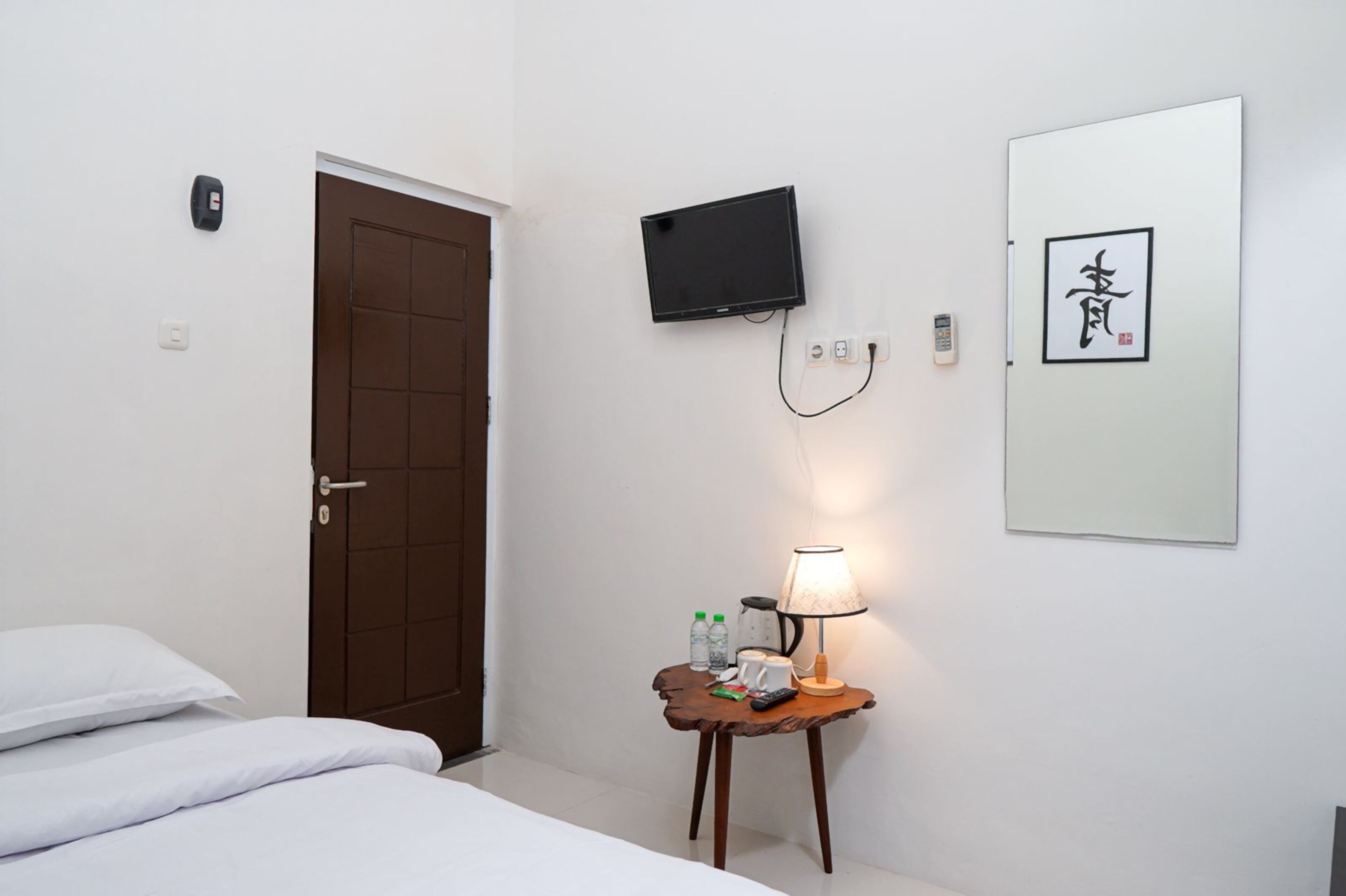 Ein Hotelzimmer mit einem Bett, einem Nachttisch mit Lampe, einem an der Wand befestigten Fernseher, einer Tür und einem gerahmten Kunstwerk.