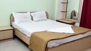 A single bed with a wooden frame, two pillows, a white bedsheet, a brown blanket, a small bedside table, and a mirror on the wall. - Vaishali Lodge (Pune)