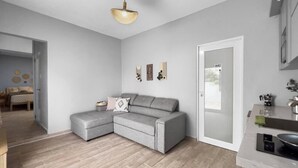 A modern living room with a grey sectional sofa, a small ottoman, and a kitchen area with a sink and countertop. - Casa La Cueva Apt 2 | Boho Oceantfront House (Arecibo)
