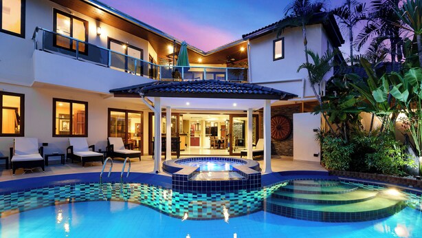 Una casa de dos pisos con piscina, mobiliario exterior y un porche cubierto.
