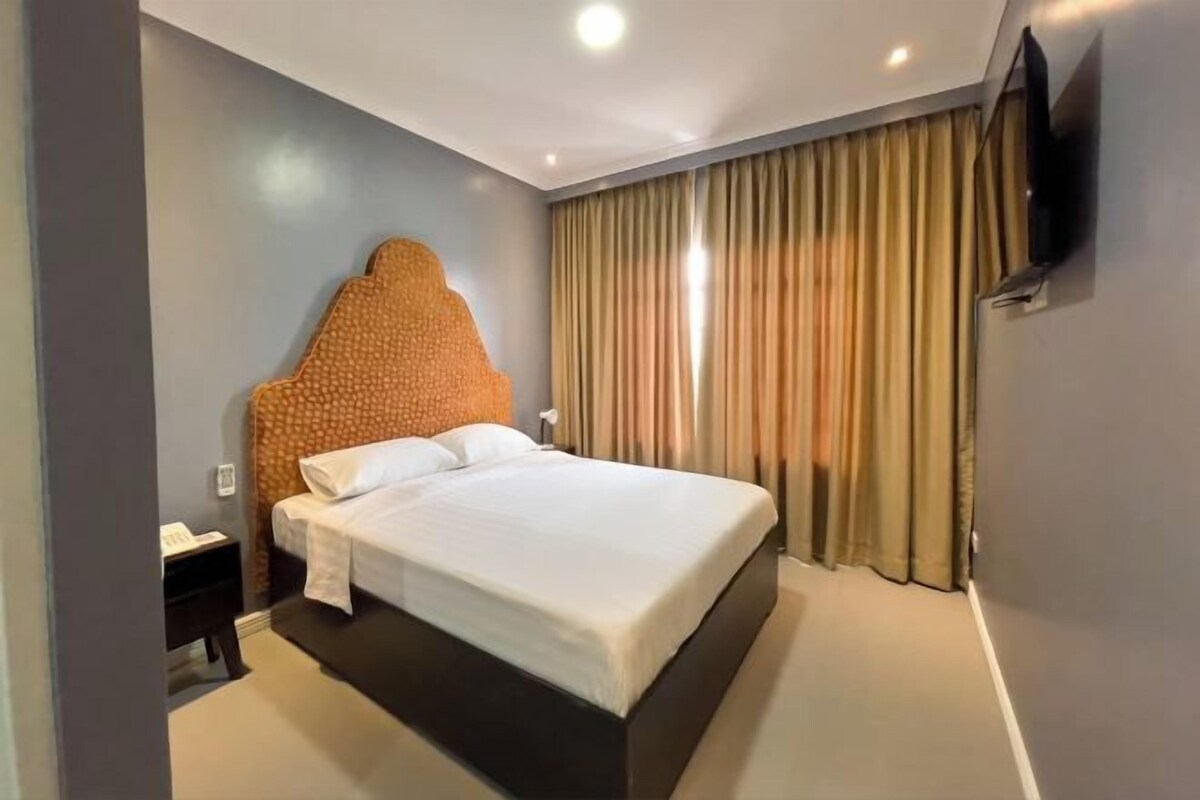 A hotel room with a large bed, a headboard, a TV mounted on the wall, and a curtain.