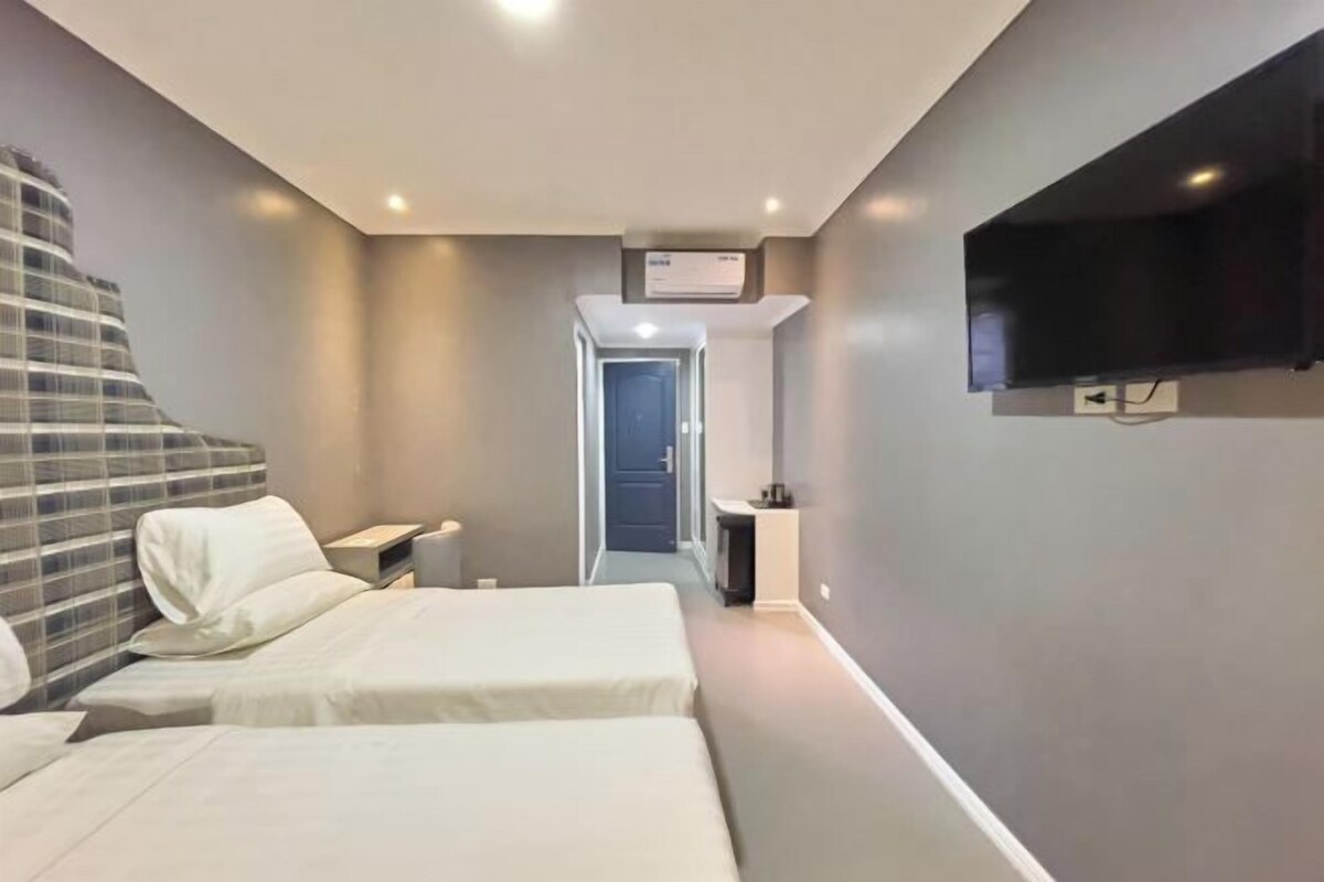 A modern hotel room with two beds, a flat-screen TV mounted on the wall, and a hallway leading to a door.