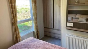 A bedroom with a bed, window with curtains, built-in wardrobe, and a radiator. - Beverley bay 2-Bed Caravan, Sleeps 6, Pet Friendly (Paignton)