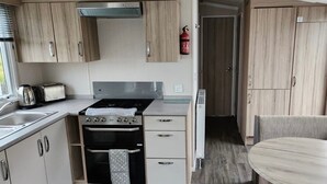 A compact kitchen with wooden cabinets, a gas stove, a built-in oven, and a small dining area with a round table and two chairs. - Beverley bay 2-Bed Caravan, Sleeps 6, Pet Friendly (Paignton)