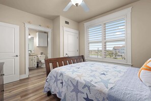 A bedroom with a bed, a window with blinds, a ceiling fan, and a view of a building outside. - Sunset Baycation - Newly Listed Next To Lazy River (Galveston)