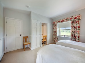 Een slaapkamer met twee bedden, een stoel, een bureau en een raam met bloemen gordijnen.