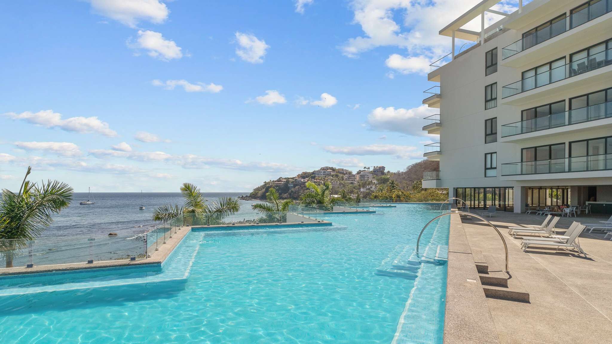 Une piscine à l’eau d’un bleu cristallin, avec vue sur la mer, et un bâtiment doté de balcons.