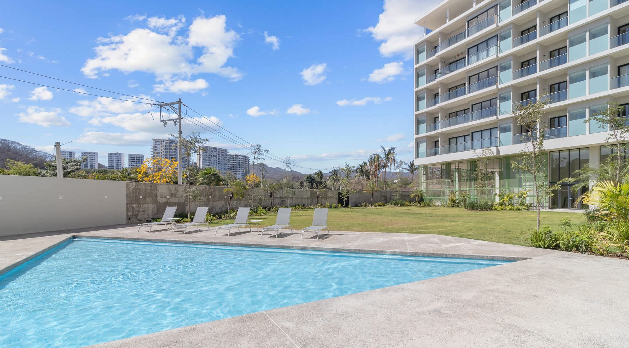 A swimming pool with lounge chairs, a modern building with balconies, and a clear sky.