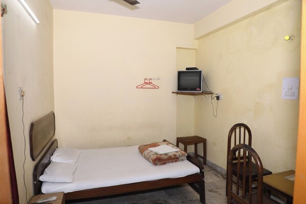 A small, simple room with a single bed, a wooden chair, a small table, and a television mounted on the wall. - Hotel Laxmi Inn (Patna)