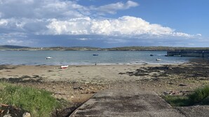 A coastal beach with boats, a rocky shore, and a distant hilly landscape under a partly cloudy sky. - NEAD NA BHFÁINLEOG (SWALLOWS’ NEST) in Claddaghduff, County Galway (Clifton)